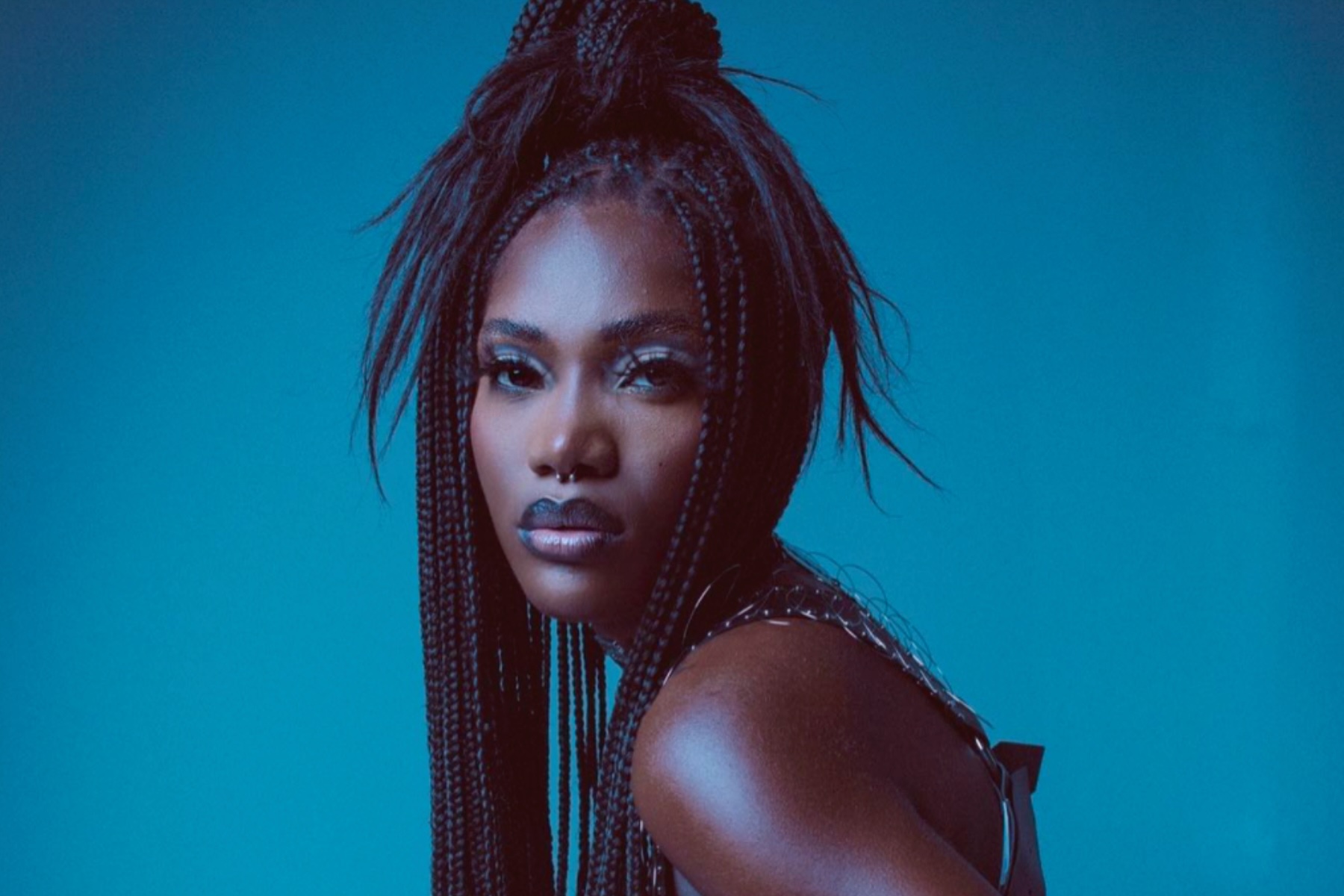 Portrait of Doechii with long braids, staring intensely into the camera, awash in blue light, on a medium blue background.